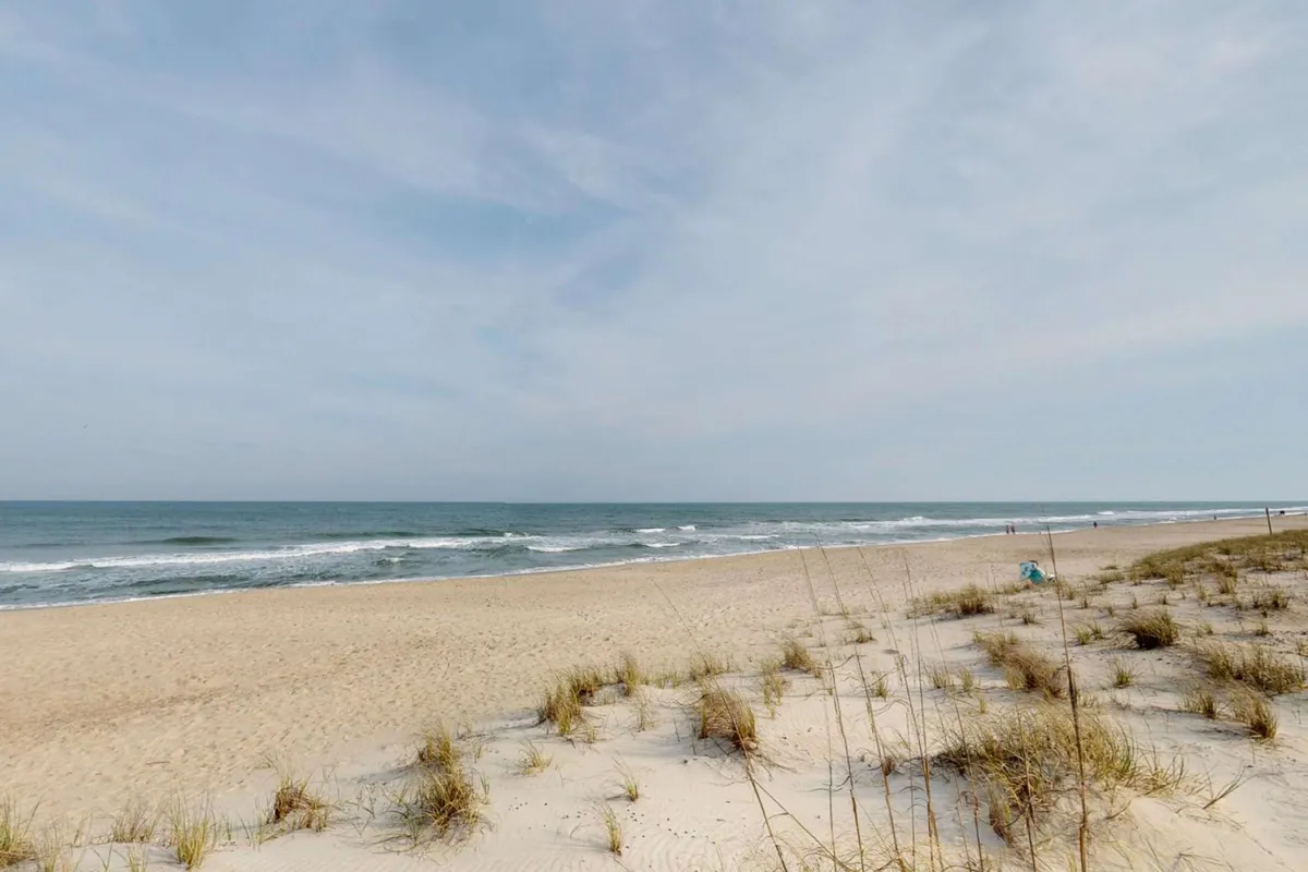 This image captures sand dunes in the foreground, gentle ocean waves, and distant beachgoers enjoying the coastline. The scene exudes a peaceful, natural seaside vibe.