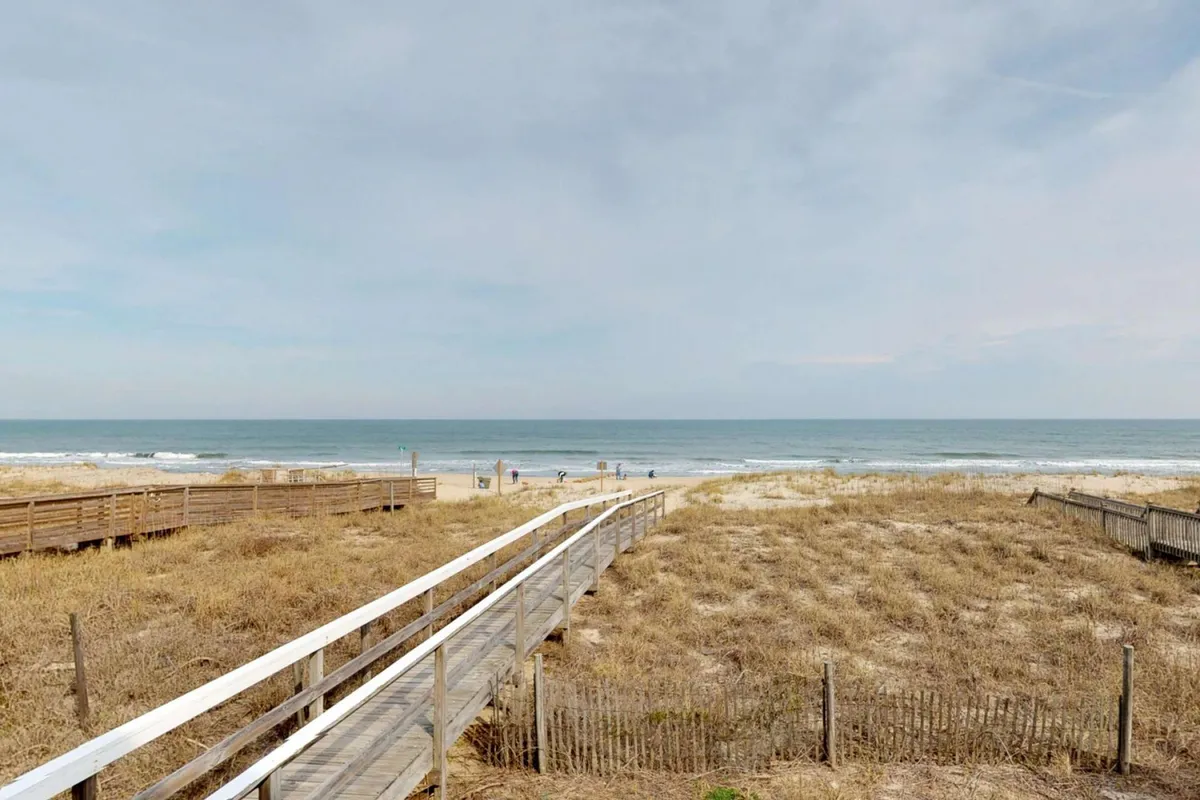 A picturesque view of a wooden boardwalk leading to the beach, bordered by dunes and the ocean beyond. Groups of people are seen strolling, suggesting easy access to local attractions and opportunities for exploration and fun moments together.