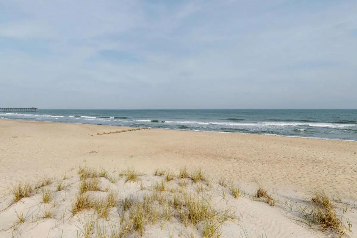 This exterior image features a sandy beach with gentle waves rolling in, dune grass in the foreground, and the iconic Kure Beach Pier visible in the distance. Families and groups can be seen enjoying the shoreline, sunbathing, and walking along the water.