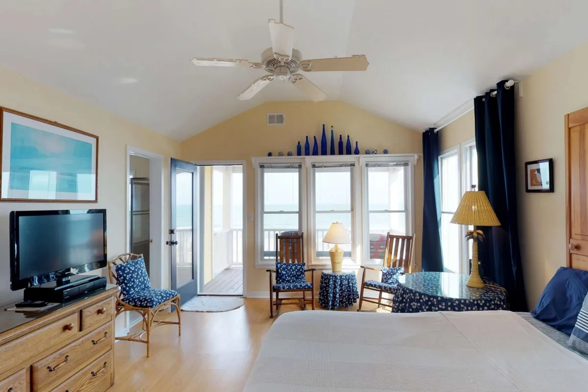 A tranquil bedroom featuring large windows with breathtaking ocean views, a bed dressed in crisp white linens, and a cozy lounge area with chairs and a table. Decorative blue glass items add a touch of coastal elegance, and the space is bathed in natural light.