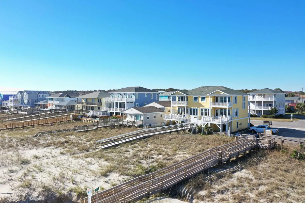 A cheerful exterior scene showcasing colorful coastal houses lining wooden boardwalks with sand dunes nearby. Children are seen playing in the surf and families gather on the beach, capturing the vibrant, welcoming atmosphere of Kure Beach.