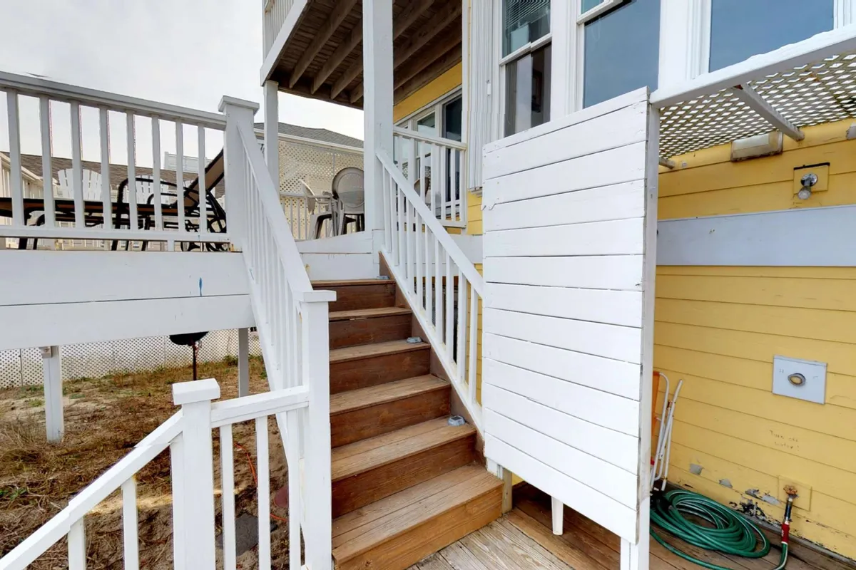 A warm, inviting exterior view of a yellow home with white wooden stairs leading to an open door, set against a backdrop of coastal greenery. The deck and stairs provide the perfect vantage point for enjoying ocean breezes, sunsets, and morning coffee.