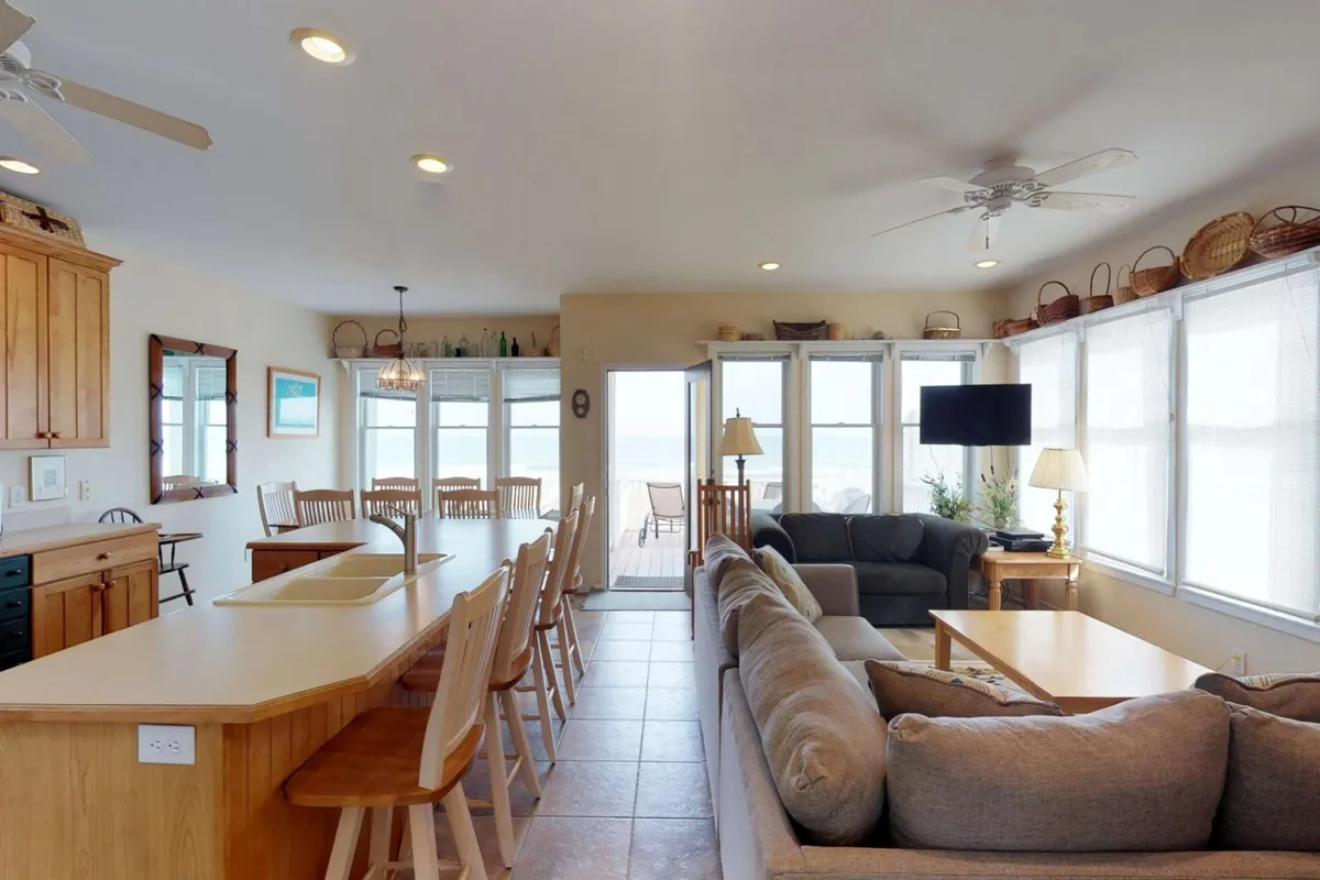 A spacious, open-concept living area featuring a kitchen island with seating, a large dining table, plush sofa, and a TV. Expansive windows showcase panoramic ocean views and fill the room with natural light. The layout encourages interaction between the kitchen, dining, and living spaces, making it ideal for group gatherings and shared activities.