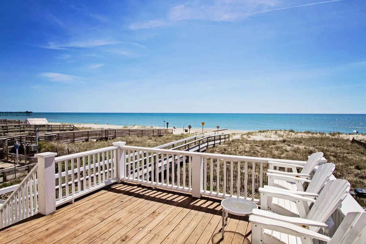 The image shows a spacious deck overlooking the Atlantic Ocean, with inviting white chairs arranged to face the water. The sturdy wooden railing frames a breathtaking, unobstructed view of the waves. The mood is tranquil, evoking a sense of relaxation and calm. The scene suggests the perfect spot for enjoying sunrises, sipping drinks, or simply unwinding while the ocean breeze drifts by.