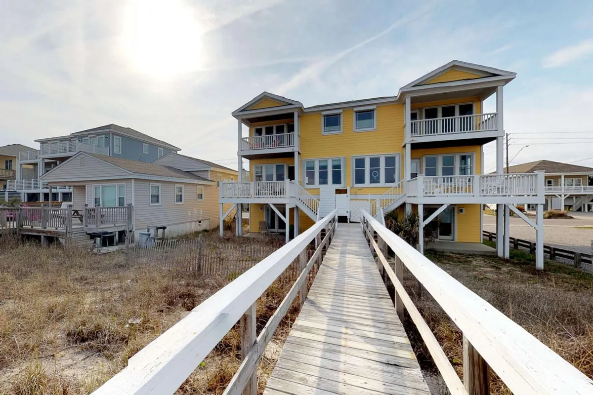 A cheerful exterior shot of the house's bright yellow facade with multiple balconies and a wooden walkway leading to the entrance. The balconies—likely some of the four decks mentioned—overlook the surrounding coastal landscape, offering hints at the various outdoor spaces available for enjoying the ocean air and views.