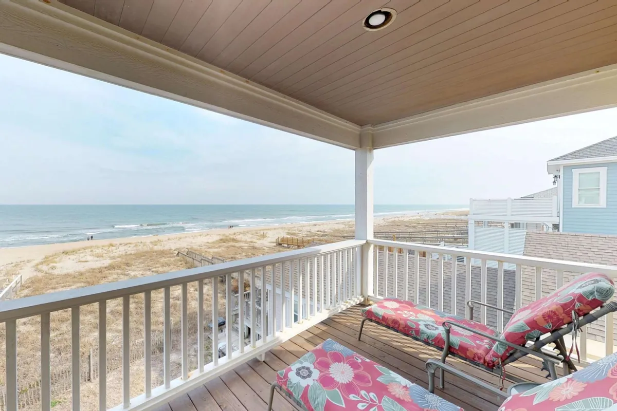 A tranquil perspective from a deck, showing lounge chairs positioned to enjoy sweeping ocean views beyond a sturdy railing. The golden sand and blue water are visible, with the gentle curve of the shore suggesting a connection to the beach below. The setup hints at relaxation, sunbathing, and quiet moments spent soaking up the coastal atmosphere.