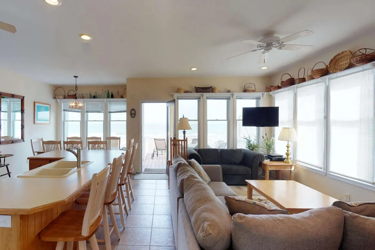 An open-plan living space with large windows letting in natural light, a comfortable seating area, a kitchen island, and a dining table set for casual meals or gatherings.