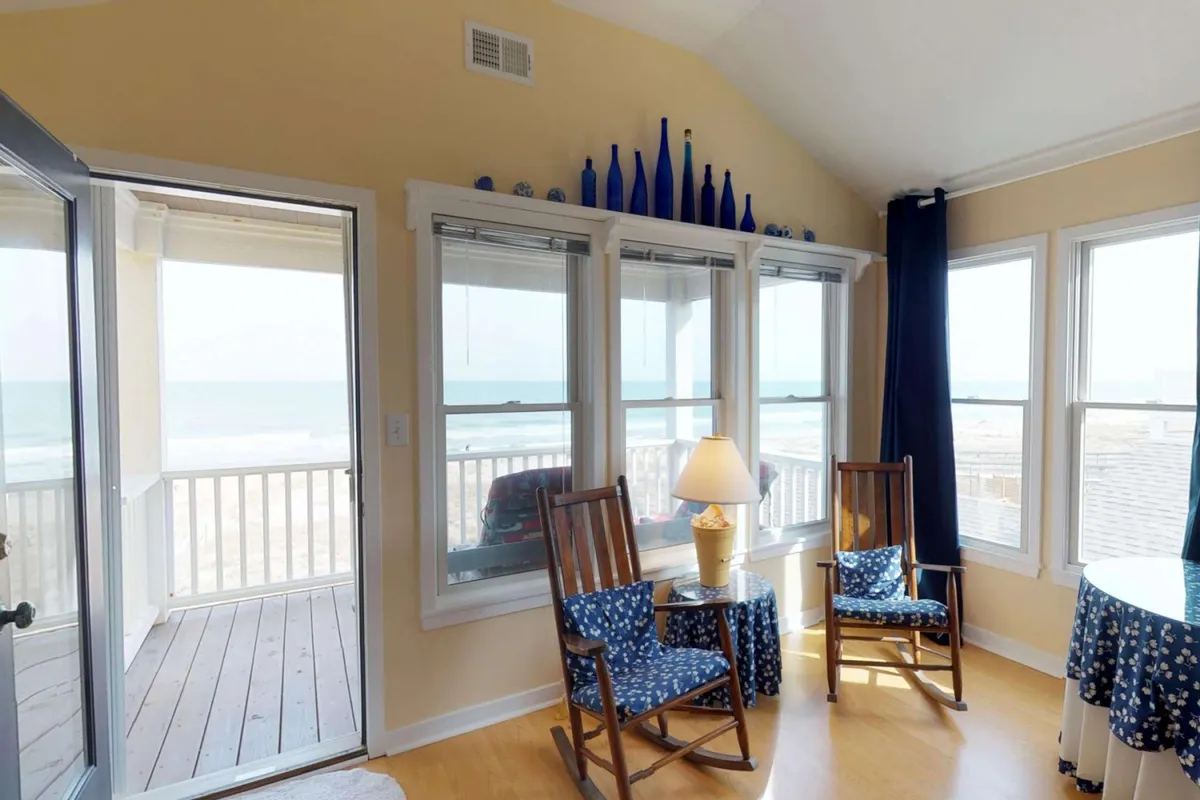 A tranquil interior scene featuring large windows that frame an expansive ocean view. Rocking chairs and blue decorative elements create a comfortable, serene spot for reading or enjoying coffee while watching the sunrise over the waves.