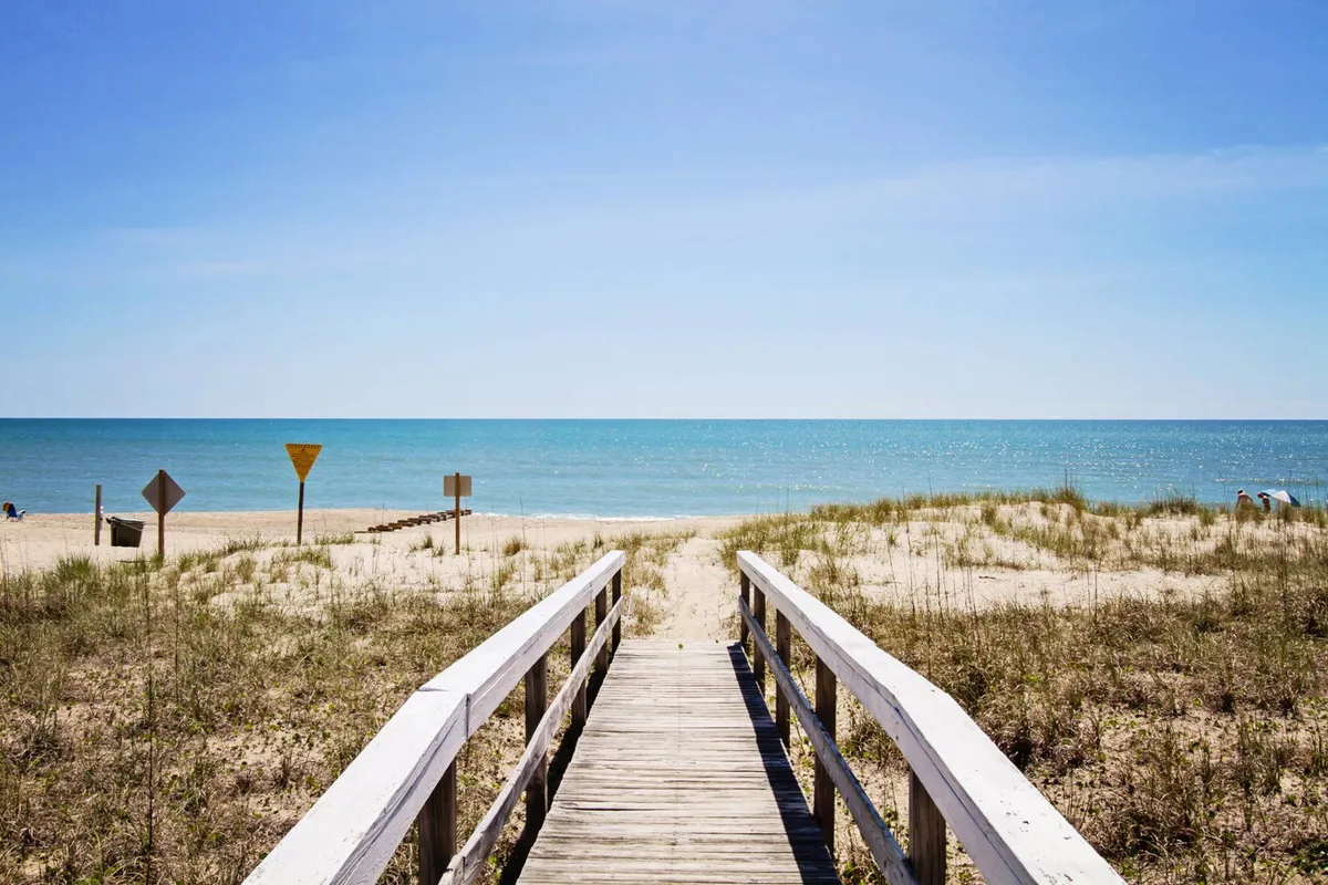 A wooden walkway extends through dune grass toward the ocean, with posted conservation warning signs visible. The beach unfolds beyond the dunes, capturing the essence of a protected coastal ecosystem. Early morning light hints at sunrise, and the area feels tranquil and undisturbed.
