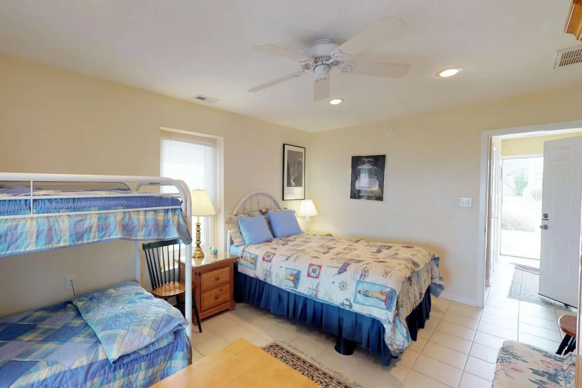 A cozy bedroom featuring both a bunk bed and a queen bed, with cheerful wall art and a nightstand with lamp. Sunlight streams in, creating a playful, family-oriented vibe ideal for kids and parents alike.